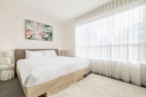 A hotel room with a large bed, a bedside table with a lamp, a wall-mounted painting, and sheer curtains. - Velvet Bloom Sunlit Comfort by Parks and Trails (Kew)