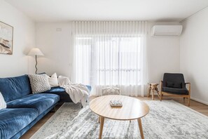 A living room with a blue sofa, a round wooden coffee table, a black armchair, and a white air conditioning unit on the wall. - Velvet Bloom Sunlit Comfort by Parks and Trails (Kew)