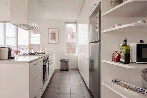 A modern kitchen with white cabinetry, a stainless steel refrigerator, a microwave, and a sink. There is a window providing natural light and a trash bin. - Garnet Gallery A Vintage Stay near Dining and Museum (Fitzroy)