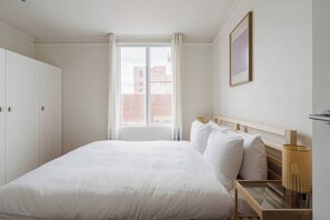 A bedroom with a large bed, a window with curtains, a wooden headboard, a wall-mounted picture, and a bedside table with a lamp. - Garnet Gallery A Vintage Stay near Dining and Museum (Fitzroy)