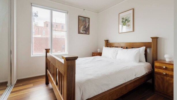 Un dormitorio con una cama de madera, ropa de cama blanca, una mesita de noche, un cuadro enmarcado y una ventana con vistas a edificios.