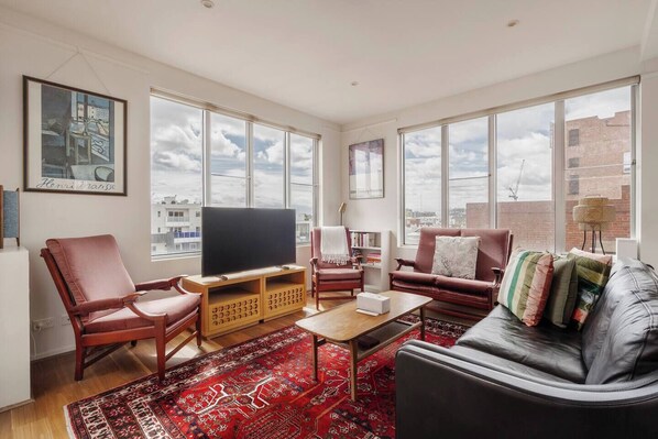 A spacious living room with a large window, a flat-screen TV, leather sofas, and a red patterned rug. - Garnet Gallery A Vintage Stay near Dining and Museum (Fitzroy)
