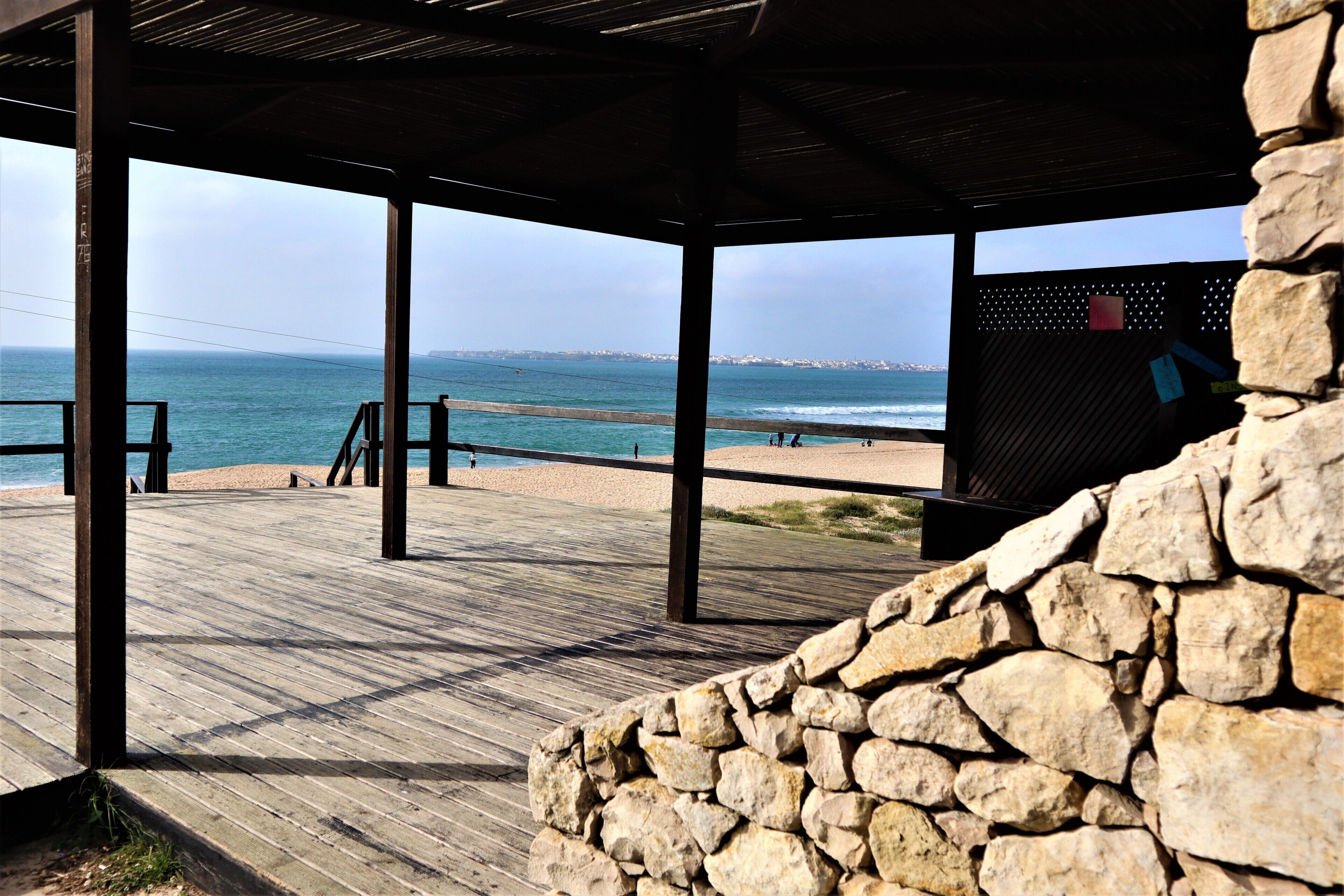 A wooden deck with a stone wall, overlooking a beach and the sea.