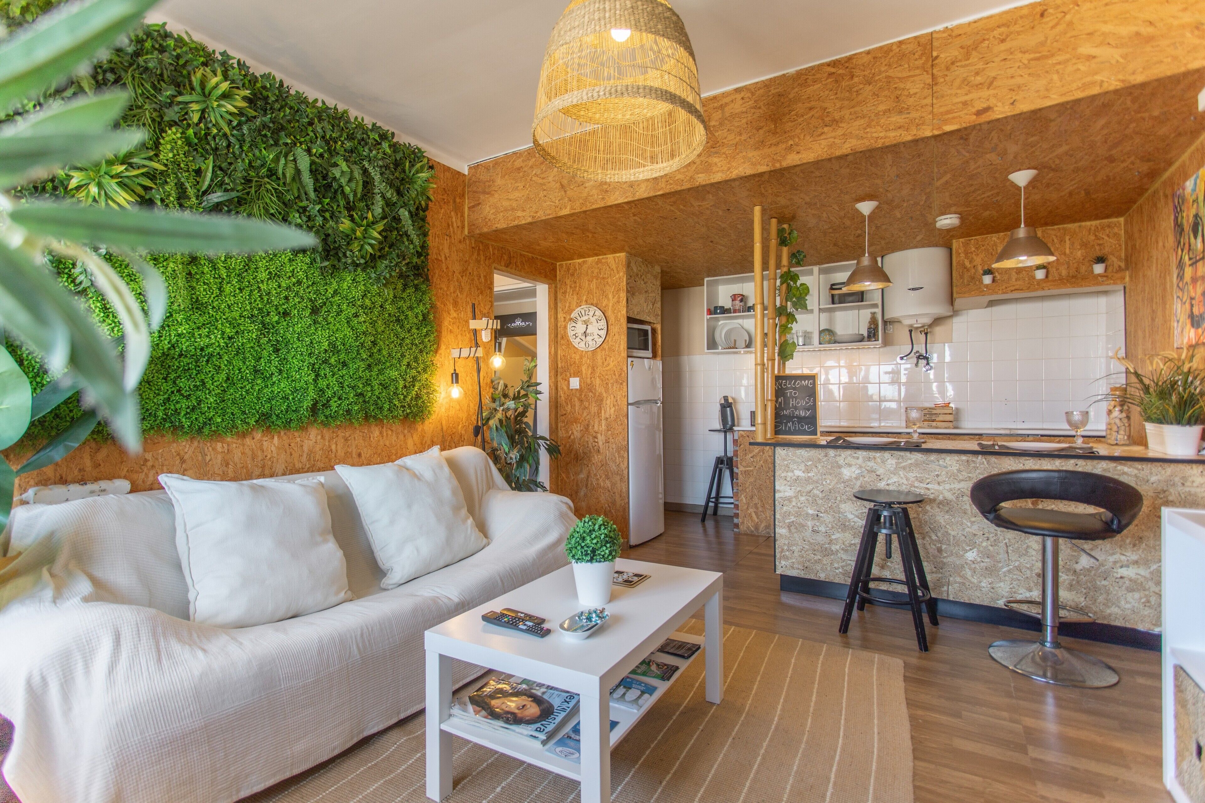 Ein modernes Wohnzimmer mit Sofa, Couchtisch und einer grünen Wand. Es gibt eine Küchenzeile mit einer Bar und Hockern.