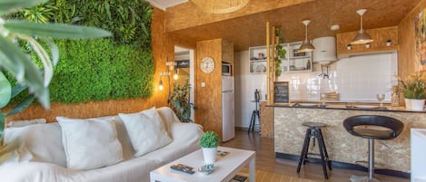 Una sala de estar moderna con sofá, mesa de centro y pared verde. Se observa una zona de cocina con barra y taburetes.