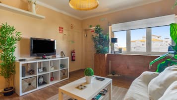 Ein Wohnzimmer mit Fernseher, einem weißen Sofa, einem Couchtisch und einem großen Fenster mit Blick auf Gebäude.