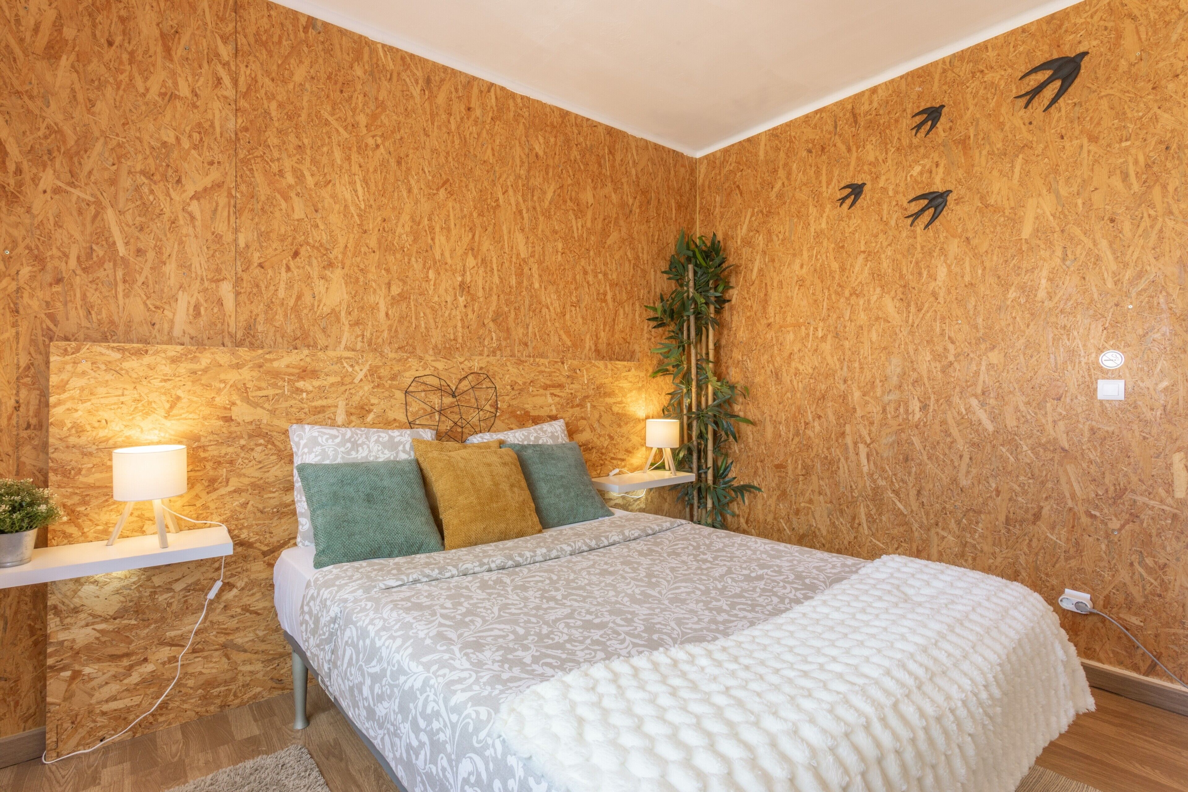 Een slaapkamer met een houten wand, een bed met beddengoed met een patroon, twee nachtkastjes met lampen, en een aan de wand bevestigde plant.