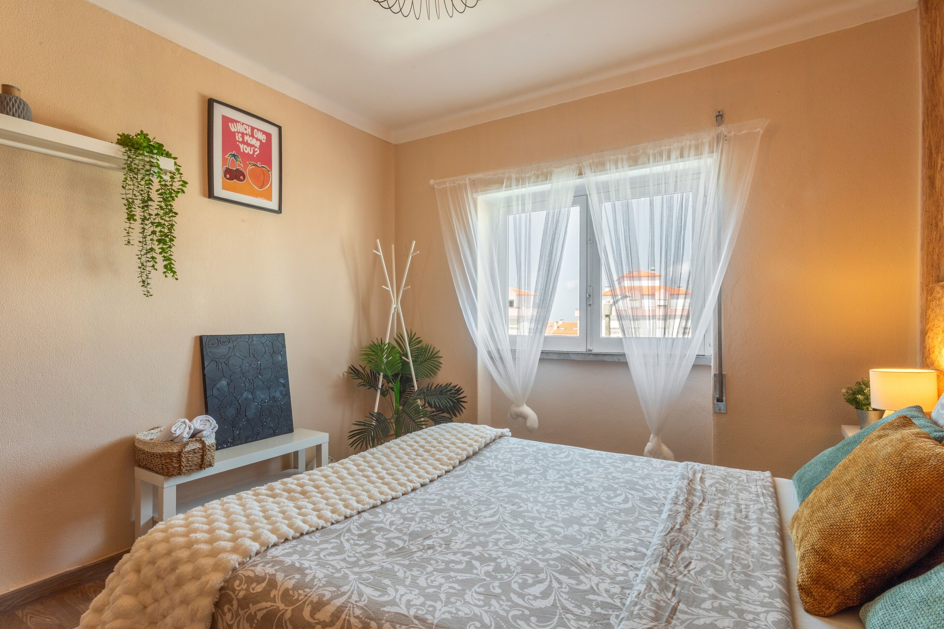 Um quarto com uma cama, uma mesinha de cabeceira, uma planta pendente, um quadro emoldurado e uma janela com cortinas leves.