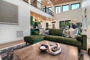 A modern living room with a green sectional sofa, a wooden coffee table, and a kitchen in the background. - Modern Cabin | Near Dollywood | Hot tub | Arcade (Sevierville)