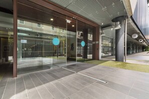 Ein moderner Gebäudeeingang mit Glastüren, Fliesenboden und einer Metallkonstruktion auf der rechten Seite.