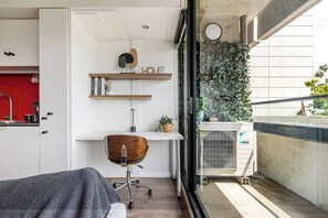 Ein kompakter Wohnraum mit Küchenzeile, einem Holzsessel, einem kleinen Schreibtisch mit Regalen und Blick auf die Stadt durch ein Fenster.