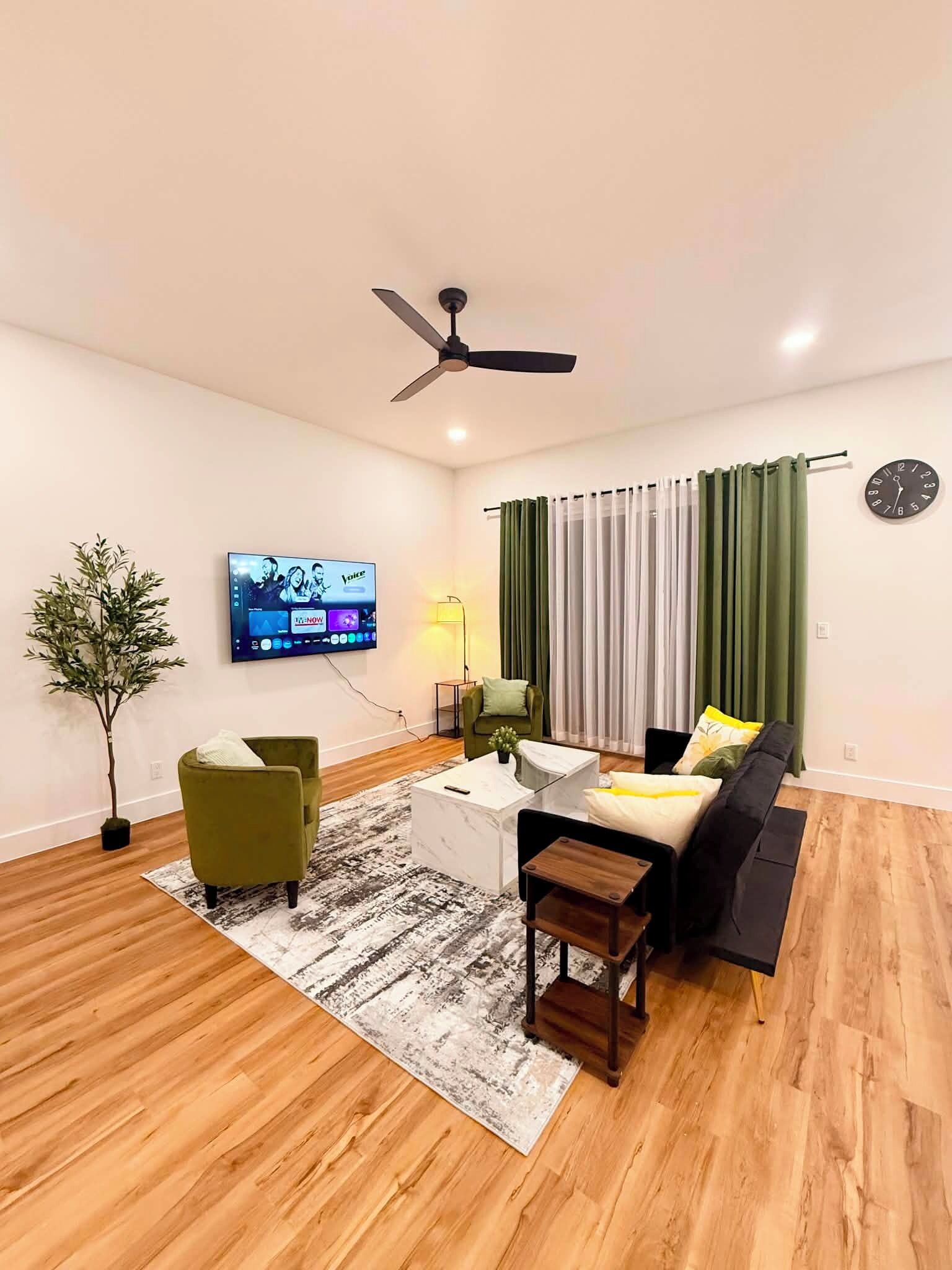 A modern living room with a flat-screen TV, a ceiling fan, a sofa, a coffee table, and a potted plant.