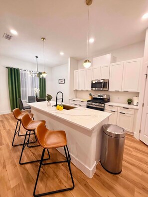 A modern kitchen with a central island, bar stools, a stainless steel sink, and a trash can. - New Built House 4 Bedroom/ 2.5 Bathroom/ 4mi to AT&T/Six Flags/Rangers (Grand Prairie)