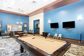 A pool table in a room with blue walls, a television mounted on the wall, and a seating area with chairs and a sofa.
