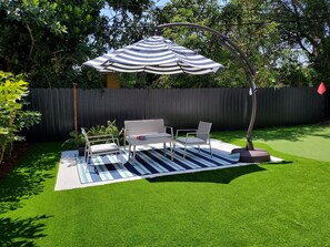 Un patio con césped artificial, un tapete exterior a rayas, una mesa metálica blanca y dos sillas, y una sombrilla a rayas.