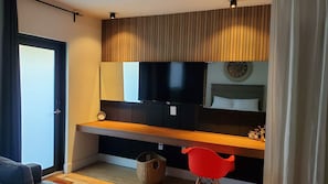 Una habitación moderna con una gran televisión de pantalla plana montada en una pared de paneles de madera, una mesita de noche con un reloj, una cama con cabecera y una silla roja.