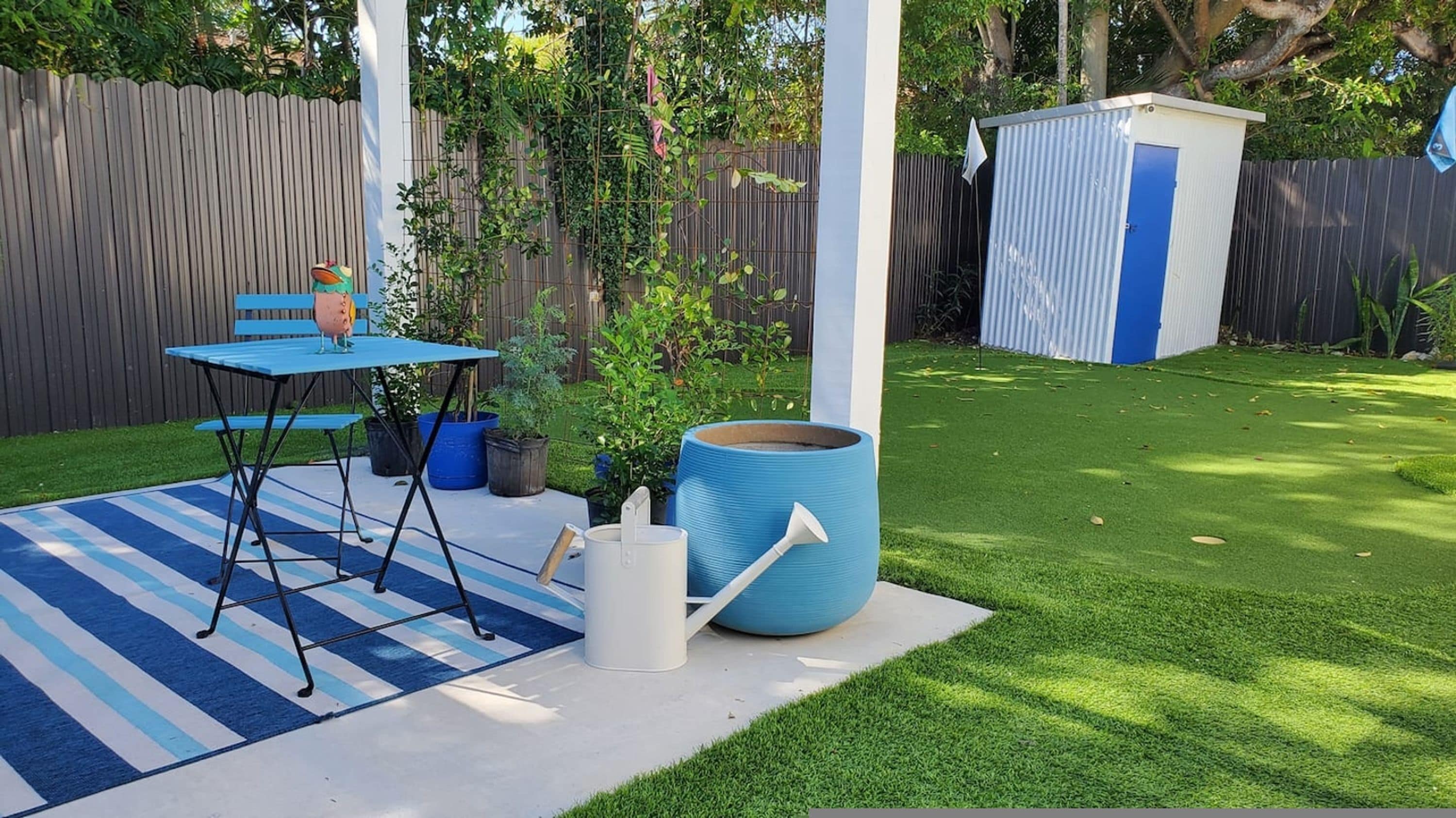 Een achtertuin met een blauwe tafel en stoelen, een grote plantenbak en een tuinhuis.