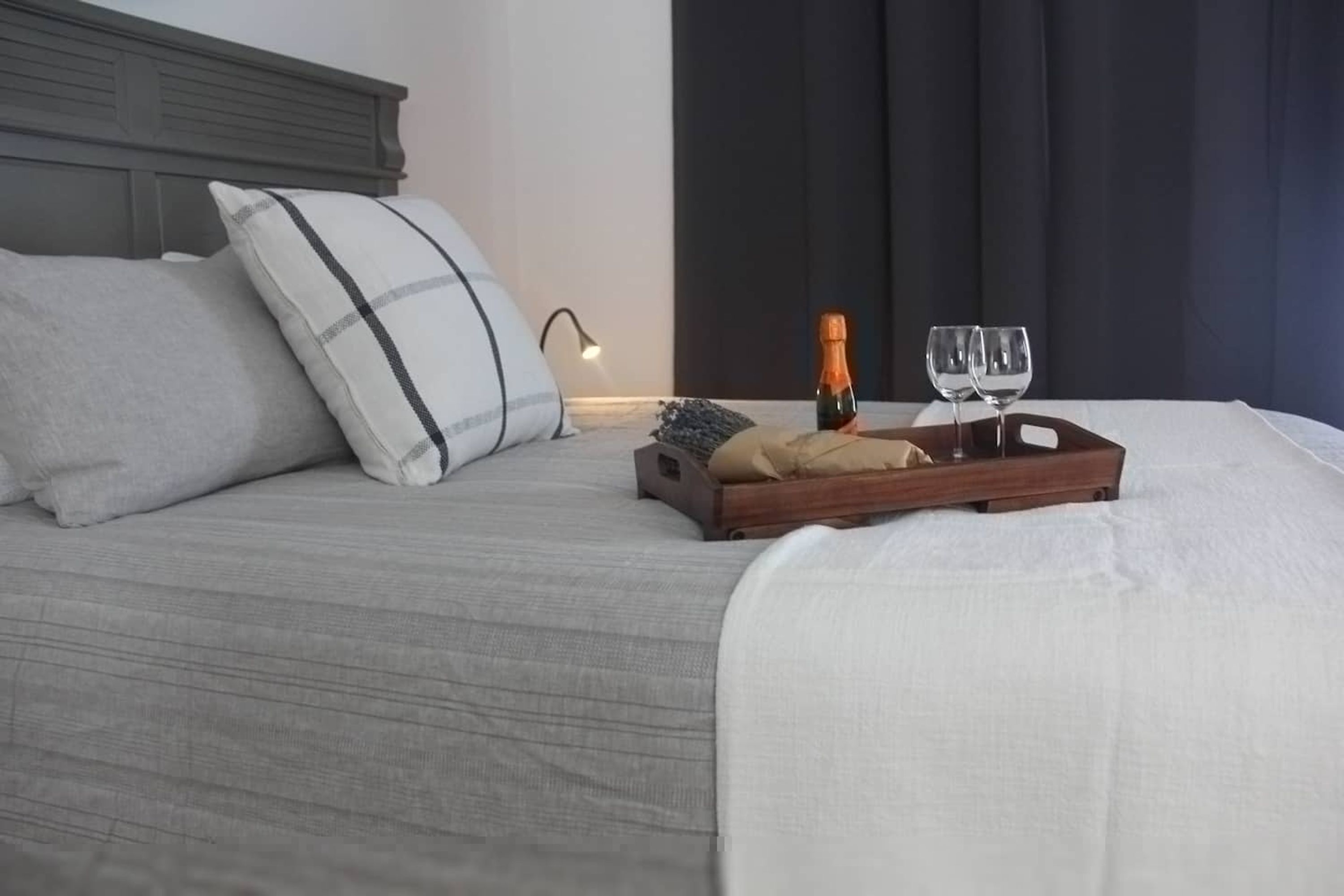 A neatly made bed with pillows, a tray with a bottle and glasses, and a bedside lamp.