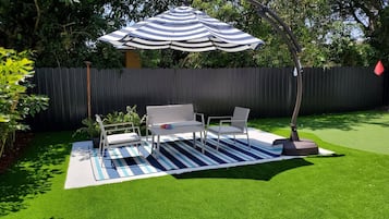 Un patio con césped artificial, un tapete exterior a rayas, una mesa metálica blanca y dos sillas, y una sombrilla a rayas.