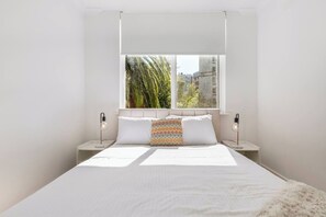 A modern bedroom with a large bed, two bedside tables, and a window with a view of palm trees and buildings. - Raleigh Escape Urban Comfort near Albert Park Lake (Windsor)