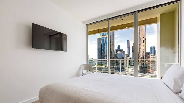 A modern hotel room with a large bed, a flat-screen TV mounted on the wall, and a city view through floor-to-ceiling windows. - Yarra Edge River and Skyline Views with Shared Pool (Melbourne)