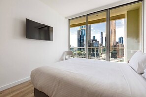 A modern hotel room with a large bed, a flat-screen TV mounted on the wall, and a city view through floor-to-ceiling windows. - Yarra Edge River and Skyline Views with Shared Pool (Melbourne)