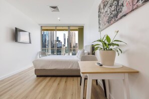 Une chambre moderne avec un lit, un bureau, une chaise, une plante en pot et une vue sur la ville à travers de grandes fenêtres.