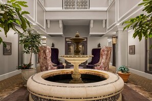 A grand hallway with a central fountain, flanked by ornate chairs and potted plants.