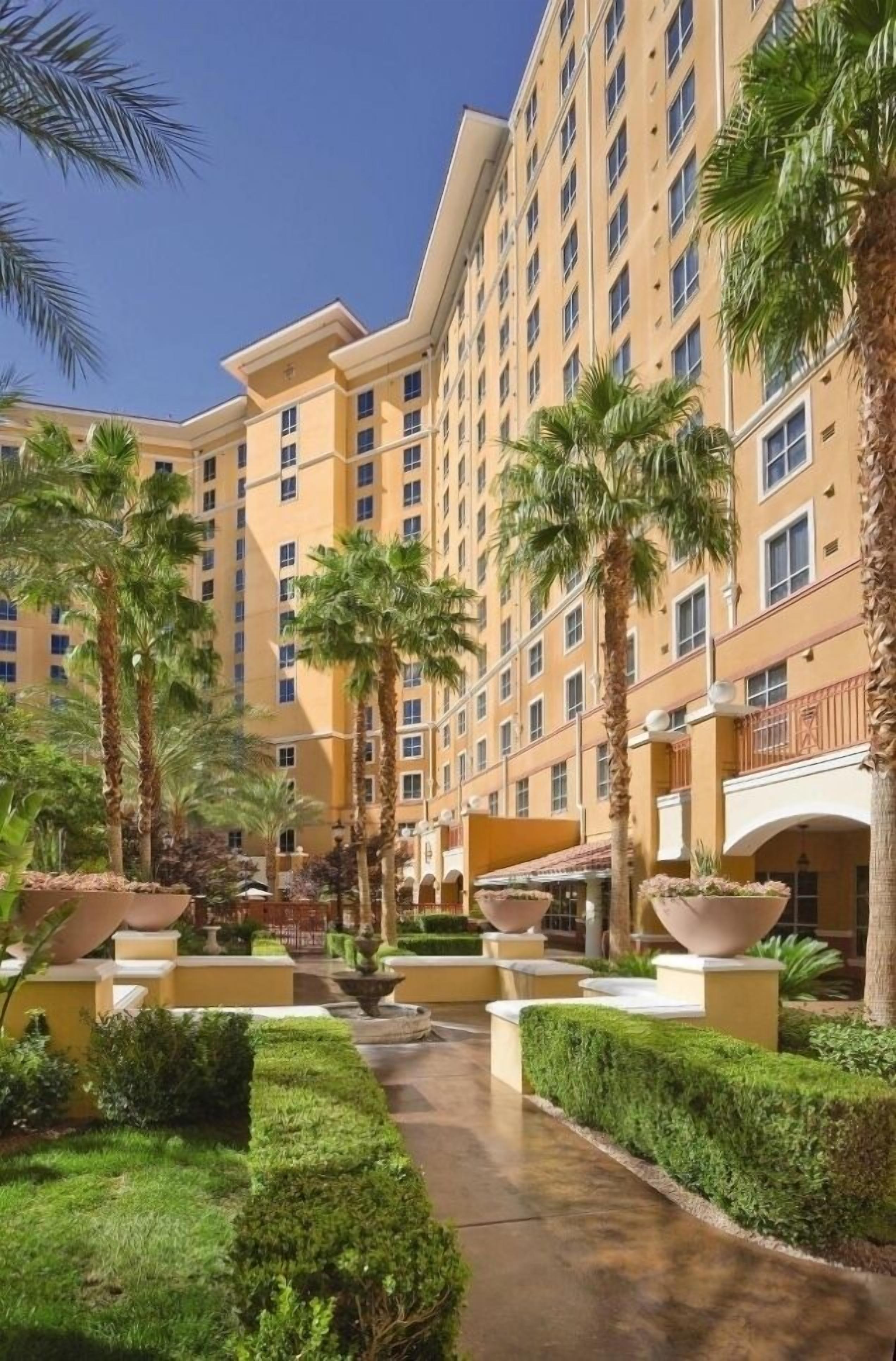A multi-story hotel with a central courtyard, palm trees, and a paved walkway.