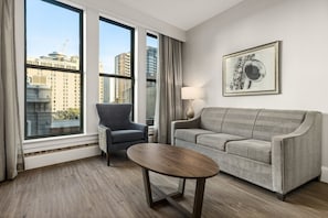 Ein modernes Wohnzimmer mit einem grauen Sofa, einem grauen Sessel, einem runden Holztisch und einer Lampe. Große Fenster bieten Blick auf die Stadtlandschaft.