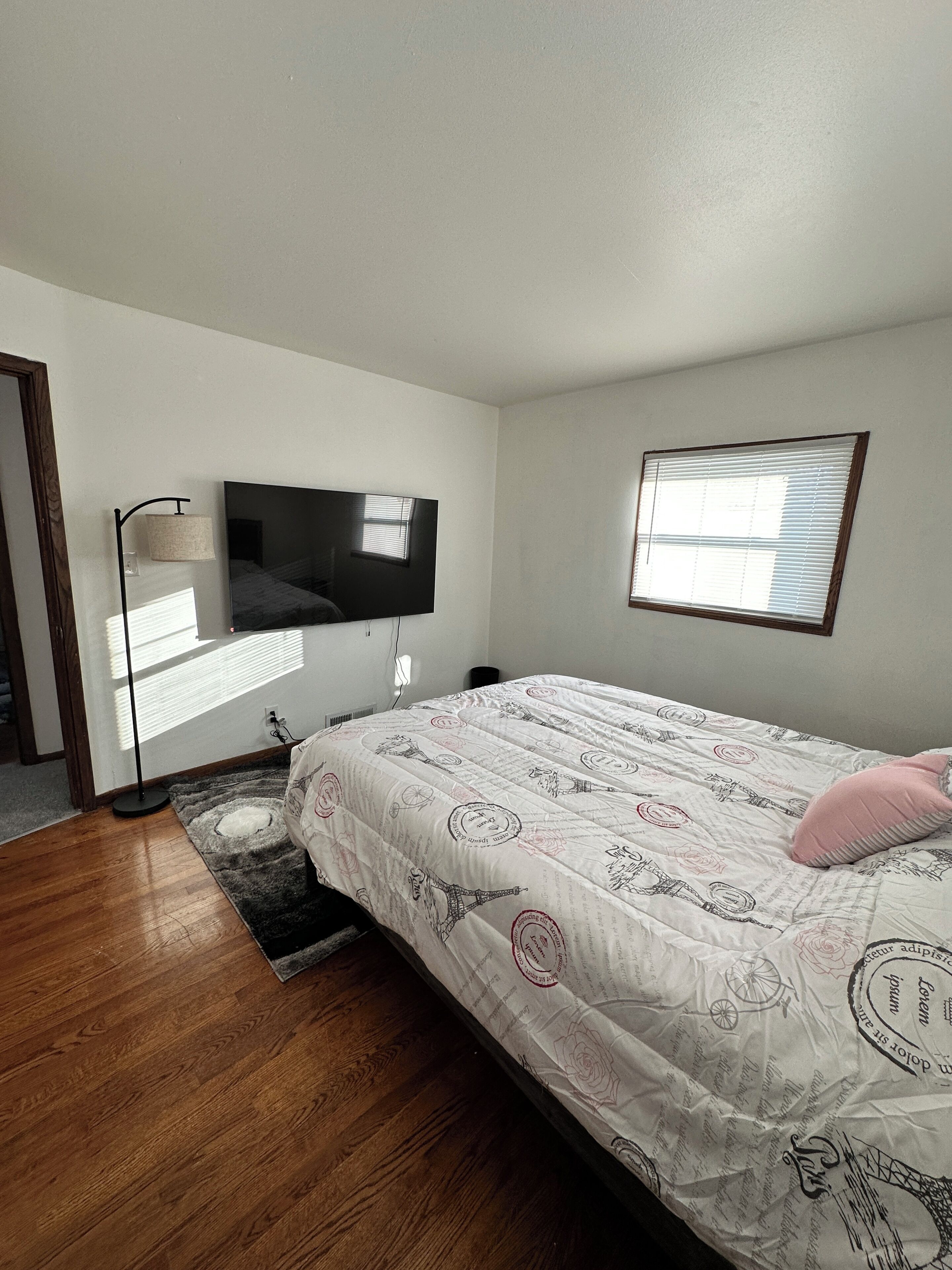 A bedroom with a bed, a television mounted on the wall, a window with blinds, and a floor lamp.