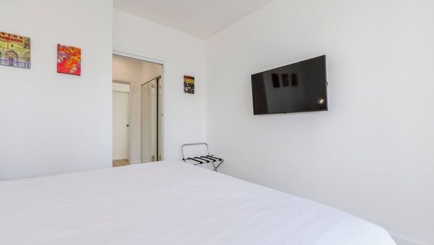 Quarto com uma cama grande, televisão fixada na parede e uma porta que dá para outro cômodo.