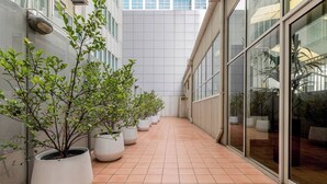 Um corredor com vasos de plantas, piso de azulejos e um edifício com grandes janelas.