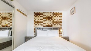 A hotel room with a large bed, a headboard with a patterned design, a bedside table with a lamp, and a coat rack. - Hampton Beach Nook Simple Stay near Dining and Beach (Hampton)