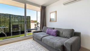 A living room with a grey sofa, a small table, and a chair. There is a window with a view of a hedge and a building. - Hampton Beach Nook Simple Stay near Dining and Beach (Hampton)