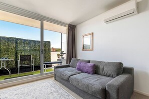 A living room with a grey sofa, a small table, and a chair. There is a window with a view of a hedge and a building. - Hampton Beach Nook Simple Stay near Dining and Beach (Hampton)