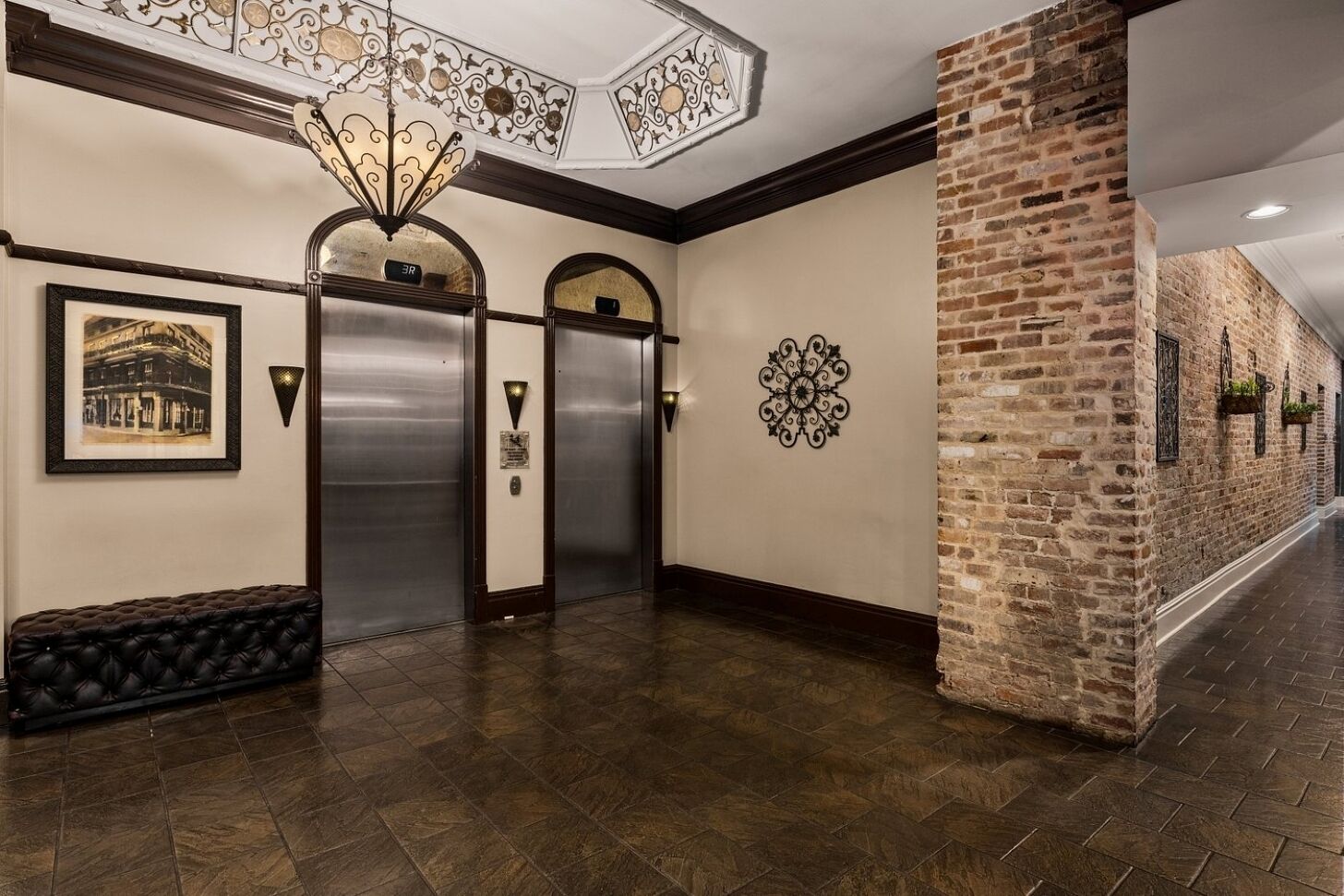 A hallway with two elevators, a decorative ceiling light, a framed picture, and a brick wall.