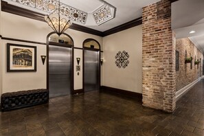 A hallway with two elevators, a decorative ceiling light, a framed picture, and a brick wall.