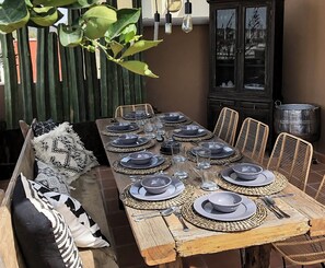 An outdoor dining area with a long wooden table set for a meal, surrounded by wicker chairs and a cushioned bench.