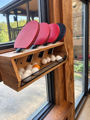Un estante de madera con paletas y pelotas de ping pong, colocado junto a una ventana que da a una terraza y follaje exterior.
