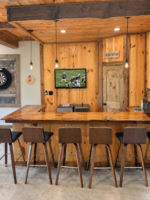 Una zona de bar con mostrador de madera, taburetes y una televisión montada en la pared, además de un dardo.