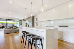 A modern kitchen with a central island, bar stools, pendant lighting, and a view of the outdoors. - The Pool Pavilion Where City meets Beach (Bentleigh)