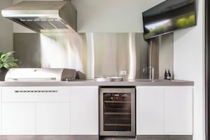 A modern kitchen with a built-in grill, a stainless steel range hood, and a flat-screen TV mounted above the counter. - The Pool Pavilion Where City meets Beach (Bentleigh)