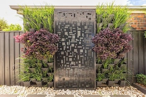 A vertical garden with a geometric metal panel, surrounded by various plants and a wooden fence. - The Pool Pavilion Where City meets Beach (Bentleigh)
