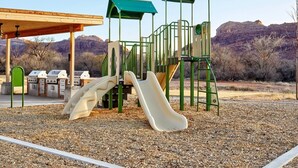 Une aire de jeux avec des toboggans et des structures d’escalade, entourée d’une zone recouverte de gravier et d’un espace barbecue.