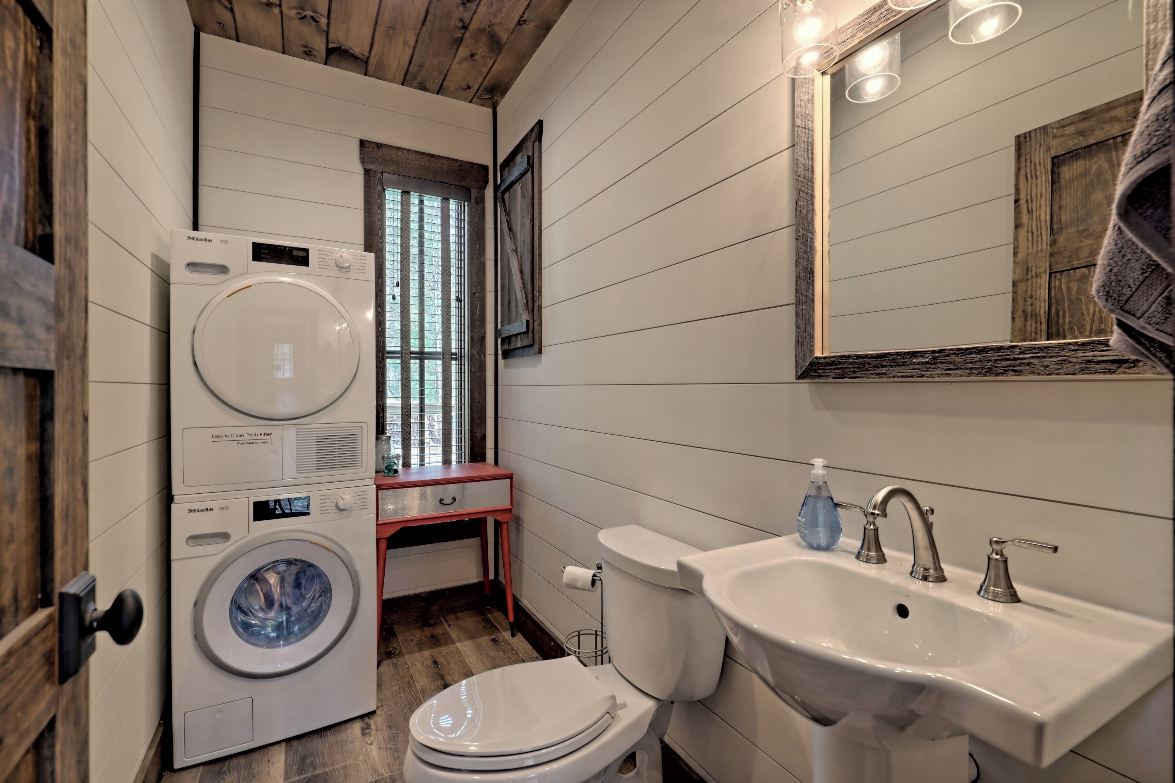 A compact bathroom with a washing machine, toilet, sink, and a window with curtains.