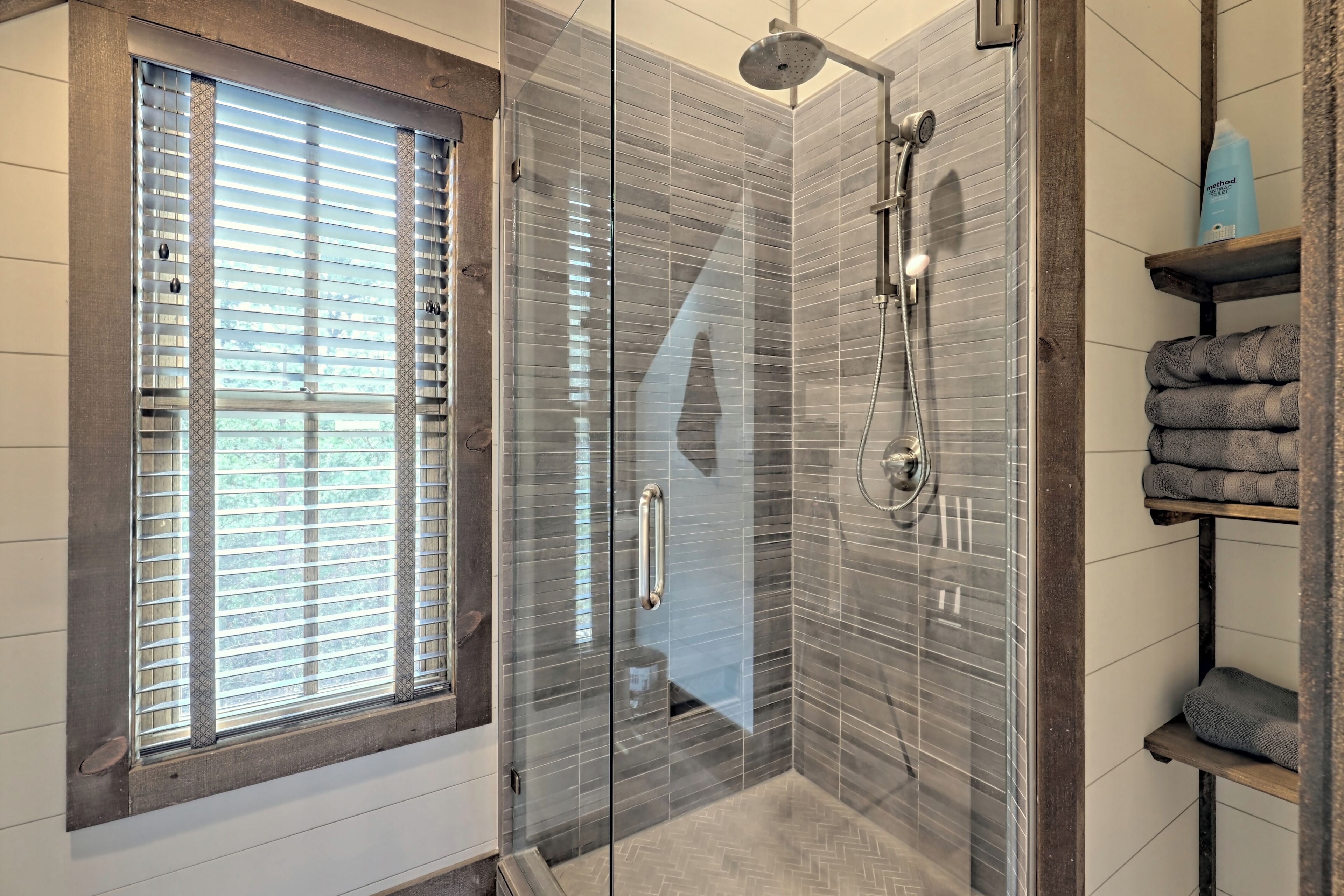 Une salle de bain moderne avec une douche à parois de verre, une fenêtre avec des stores et un porte-serviettes sur lequel sont pliées des serviettes.