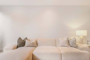 Un soggiorno moderno con un divano angolare beige, diversi cuscini e un comodino con una lampada.