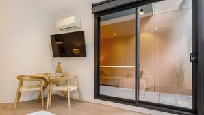 A modern living room with a flat-screen TV mounted on the wall, a wooden dining table with chairs, and a sliding glass door leading to a balcony with a sofa and a plant. - Mariner Resort Style Escape Where City meets Coast (Port Melbourne)