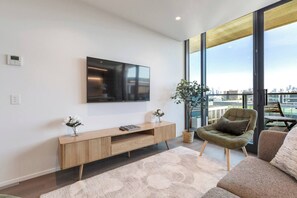 A modern living room with a flat-screen TV, a wooden console table, a potted plant, and a view of the city through large windows. - The Parkhouse Riverside Living with Pool and Sauna (Abbotsford)